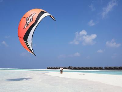 Personne en kitesurf au-dessus d'une plage de sable blanc et d'eau turquoise sous un ciel bleu.