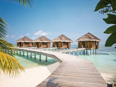 Holzhäuschen auf Stelzen mit Reetdächern über klarem, türkisfarbenem Wasser, verbunden durch Steg.