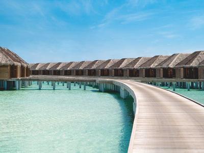 Ein Steg führt über türkisfarbenes Wasser zu mehreren über dem Wasser gebauten Hütten mit Strohdächern.