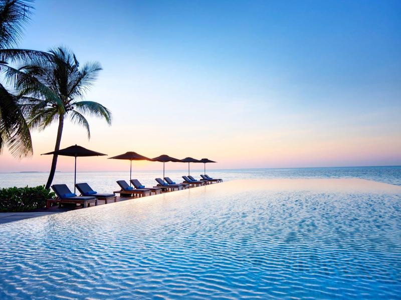 Infinity-Pool mit Liegestühlen und Sonnenschirmen, Palmen, ruhigem Wasser und blauem Himmel im Sonnenuntergang.