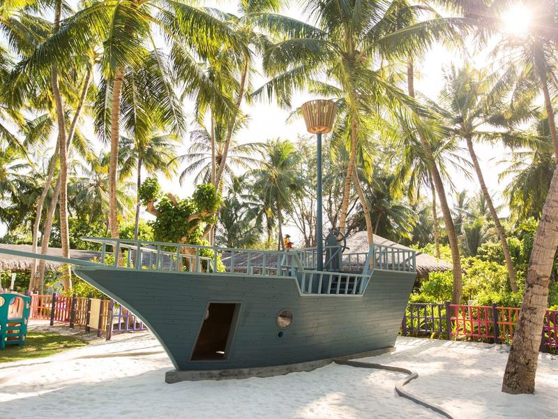 Graues Holzschiff-Spielgerät im Sand, umgeben von hohen Palmen und Sonnenlicht.