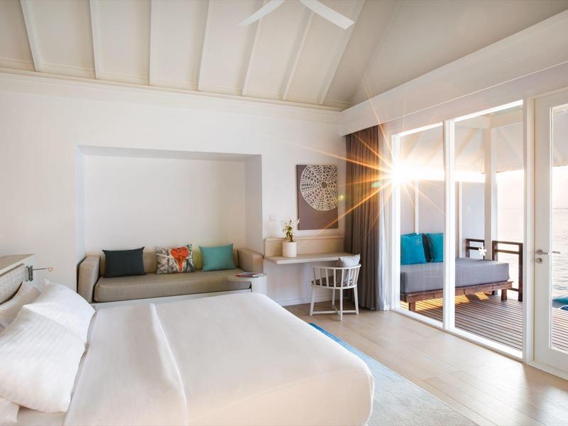 Weißes Schlafzimmer mit Doppelbett, grauem Sofa, Schreibtisch und drei großen Spiegeln an der Wand.