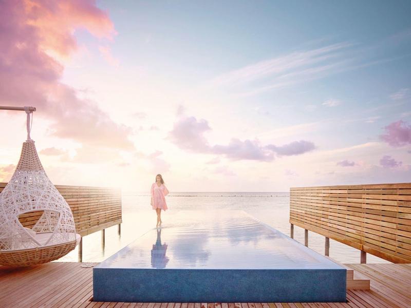 Person steht am Infinity-Pool mit Blick auf ruhiges Meer, Pastellhimmel und hölzerne Umzäunung.