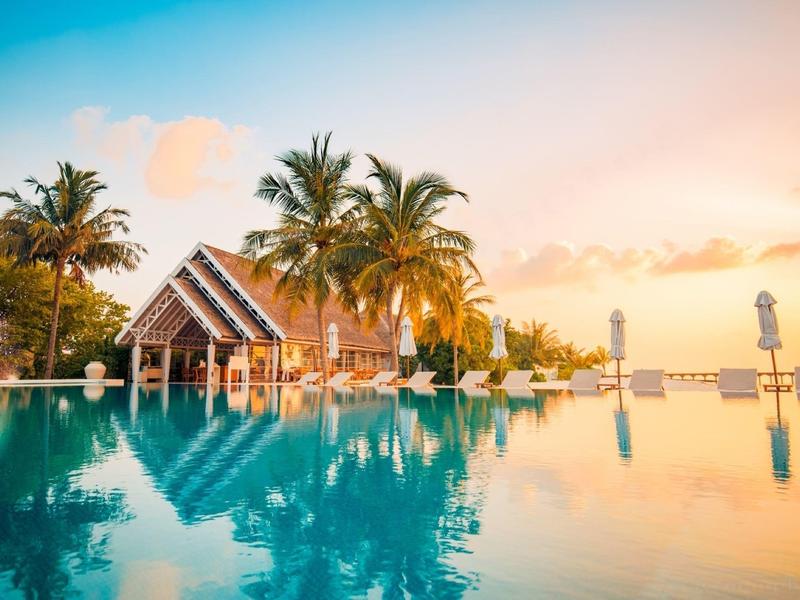 Infinity-Pool mit Palmen und Holzhaus bei Sonnenuntergang am tropischen Urlaubsort.