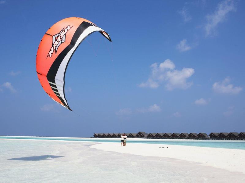 Ein roter Kitesurf-Schirm fliegt über einem weißen Sandstrand und türkisfarbenen Wasser bei blauem Himmel.
