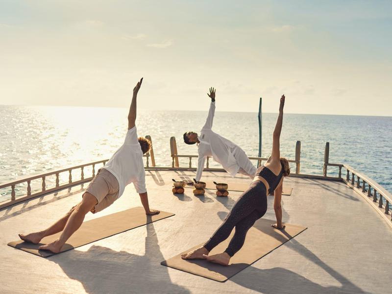 Vier Menschen machen Yoga in Seitstützposition auf einer Bootsterrasse im Sonnenlicht am Meer.