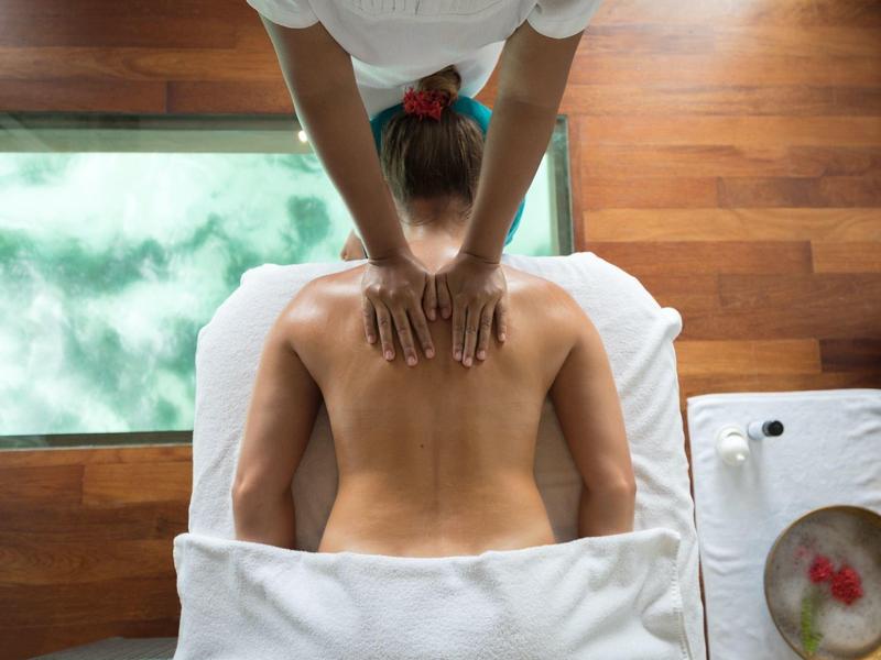 Person liegt auf Massagebank, wird von Therapeut mit Händen am Rücken massiert, Holzfußboden sichtbar.