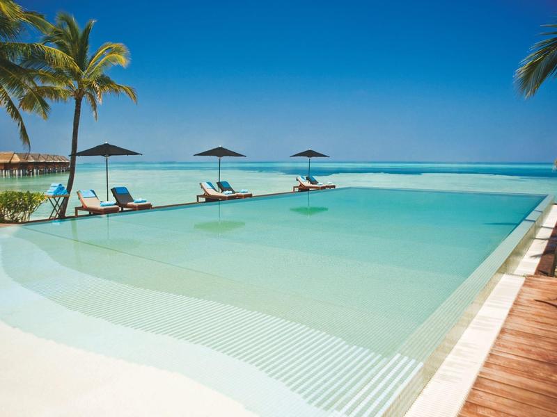 Großzügiger Infinity-Pool mit Liegestühlen und Sonnenschirmen, Palmen und blauem Himmel am Meer.