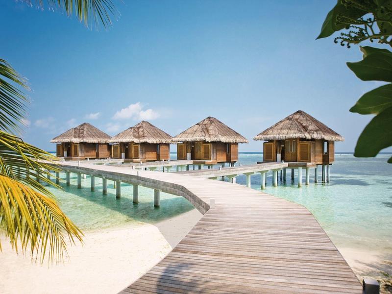 Holzhäuschen auf Stelzen mit Reetdächern über klarem, türkisfarbenem Wasser, verbunden durch Steg.