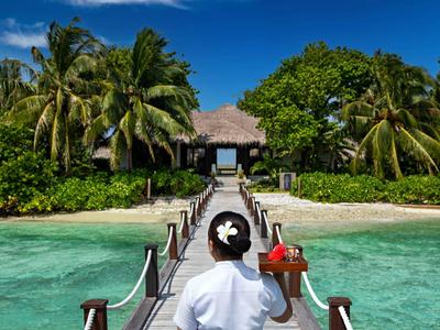 Ein Hotelangestellter trägt ein Tablett auf einem Steg zum Inselresort mit tropischer Vegetation.