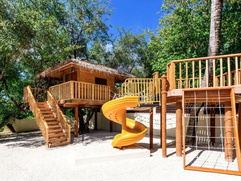 Ein Baumhaus mit Spielplatz und Rutsche in einer sonnigen, bewaldeten Umgebung.