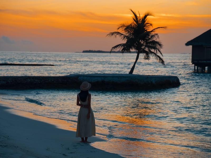 Person steht am Strand bei Sonnenuntergang mit Palmen und Überwasserbungalow im Hintergrund.