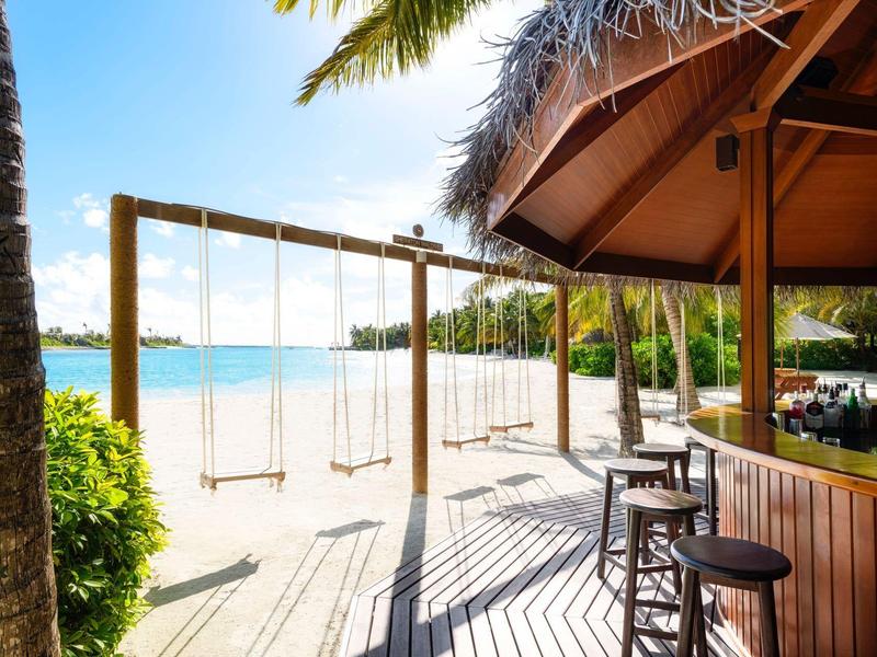 Strandbar mit Hockern und Schaukelgerüst vor blauem Meer unter klarem Himmel