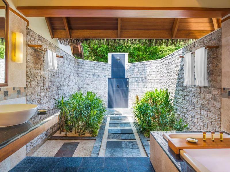 Freiluftbad mit Badewanne und Waschbecken, umgeben von Steinmauern und Pflanzen.