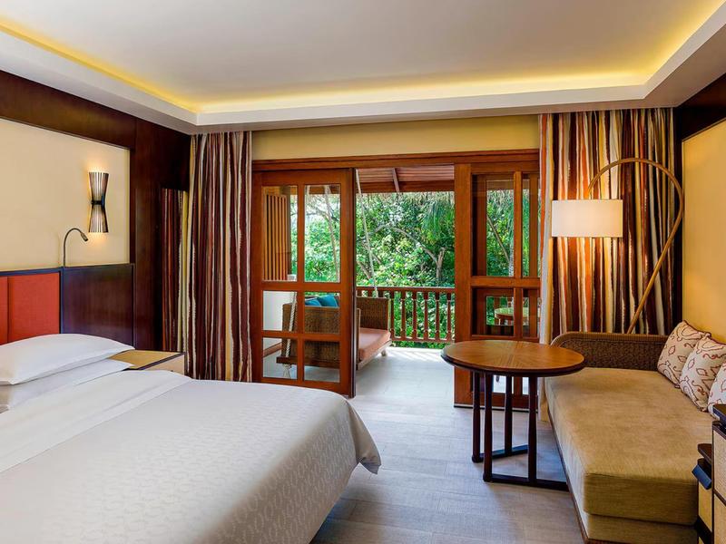 Hotelzimmer mit Doppelbett, Sofa, Tisch und Balkon mit Blick auf grüne Bäume.