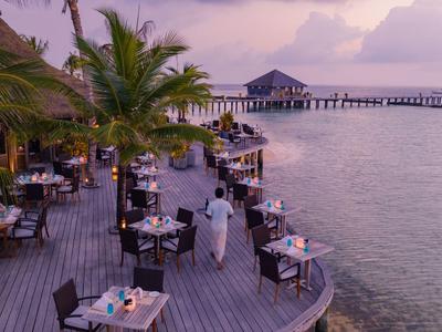 Offenes Restaurant am Meer mit Holzboden, Tischen, Stühlen und Palmen bei Sonnenuntergang.