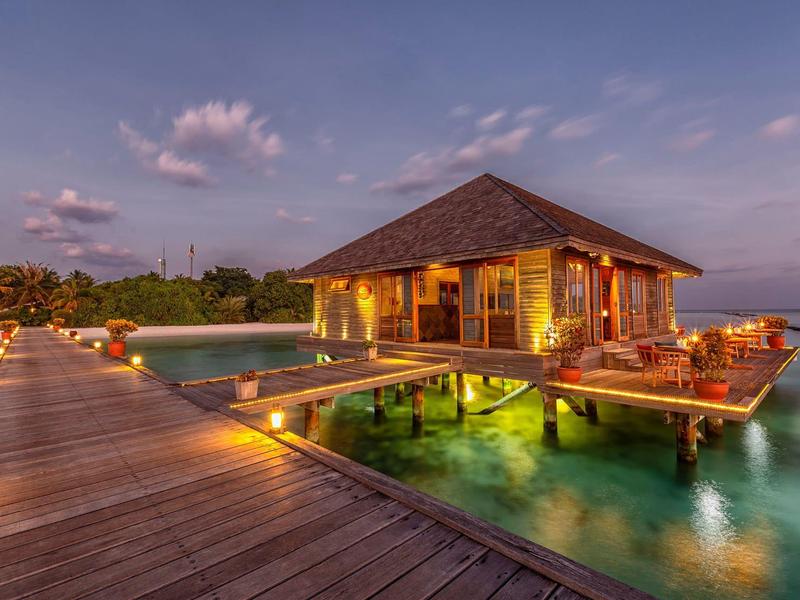 Über Wasser gelegenes Ferienhaus mit Holzsteg und beleuchtetem Pool bei Sonnenuntergang.