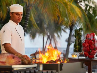 Homme en tenue de chef blanc préparant des grillades dans un cadre tropical avec des palmes.