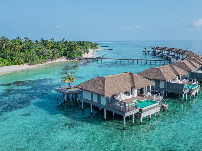 Überwassertbungalows mit Stroh­dächern auf türkis­farbenem Wasser, grüne Insel im Hintergrund.