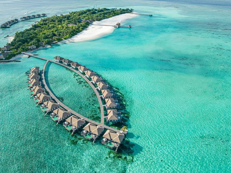 Tropische Insel mit türkisfarbenem Wasser und Bungalows auf Stelzen, die ins Meer ragen.