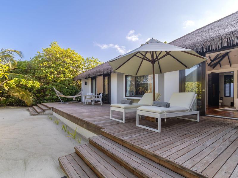 Modernes Strandhaus mit Holzterrasse, Sonnenschirm und weißen Loungemöbeln vor grünem Buschwerk.