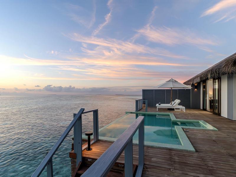 Holzterrasse mit Pool, Liegestühlen und Blick auf ruhiges Meer bei Sonnenuntergang.