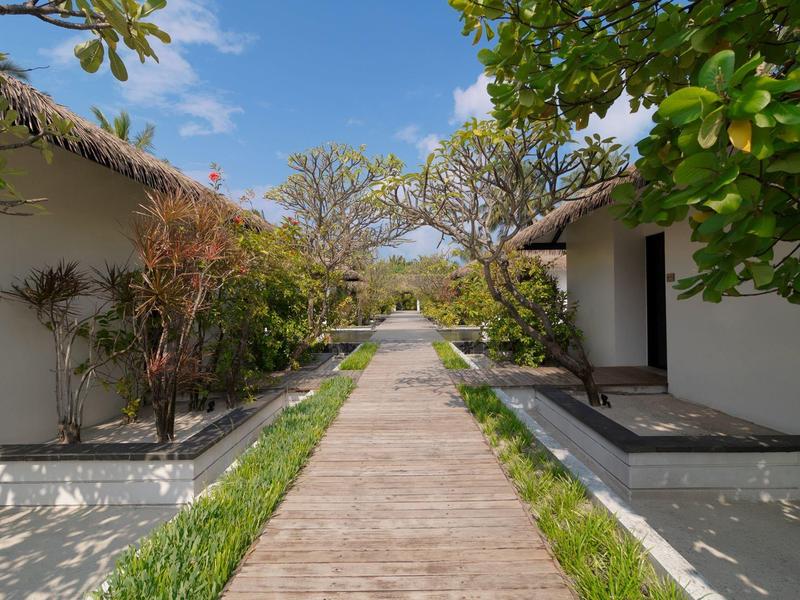 Holzweg zwischen kleinen weißen Bungalows, umgeben von grünem Laub und blauem Himmel.