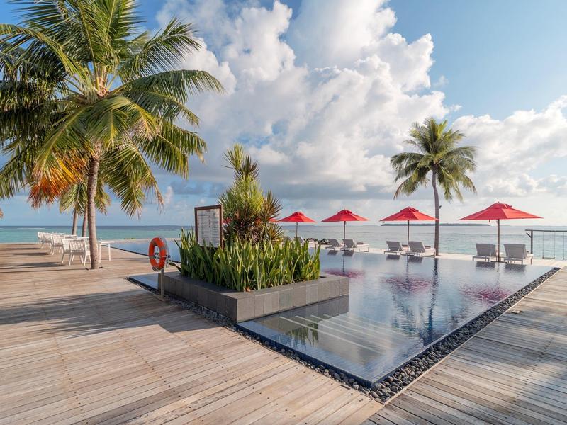 Moderner Infinity-Pool mit Holzterrasse, Palmen und roten Sonnenschirmen am Meer unter blauem Himmel.