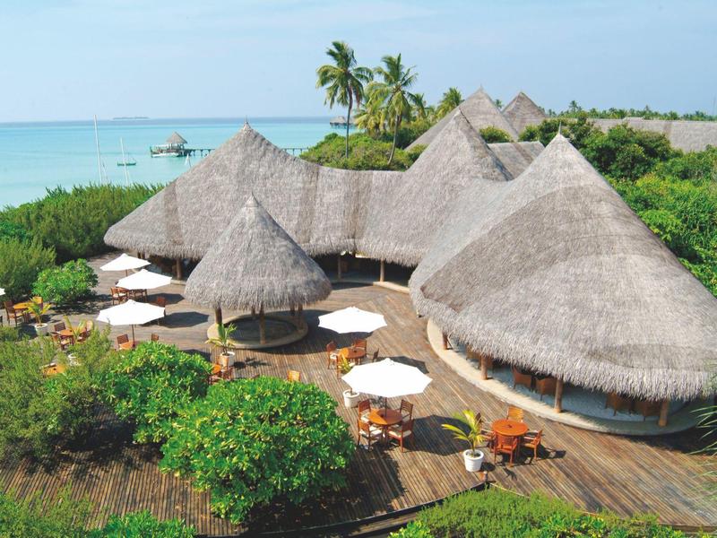 Reetgedeckte Bungalows am Strand mit weißen Sonnenschirmen und Palmen im Hintergrund.