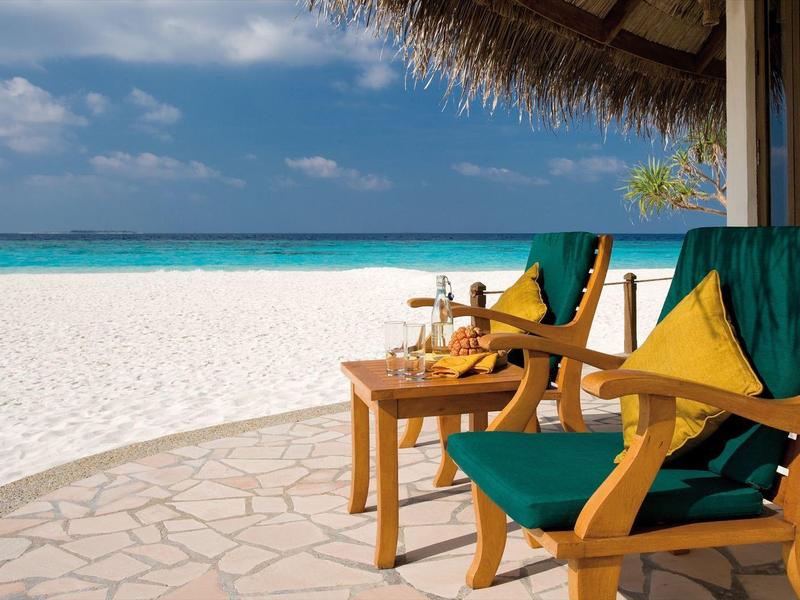 Zwei Holzstühle mit grünen Kissen auf einer Terrasse mit Blick auf einen weißen Sandstrand und das blaue Meer.