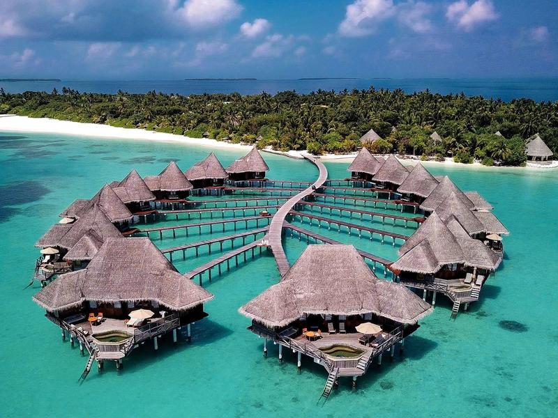 Luxuriöse Wasserbungalows mit Strohdächern über klarem, türkisfarbenem Meerwasser vor einer tropischen Insel.
