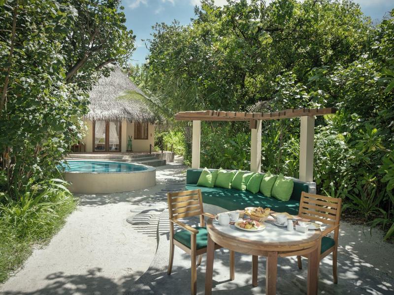 Ein runder Tisch mit Stühlen und Getränken vor einem Pool und einem strohgedeckten Bungalow, umgeben von tropischer Vegetation.