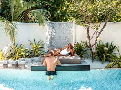 Zwei Personen entspannen am Rand eines blauen Pools mit Palmen und grünen Pflanzen dahinter.