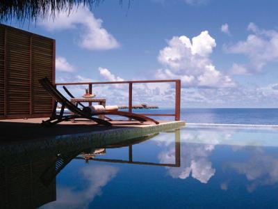 Pool mit Wasser, Liegestühle und blauer Himmel mit Wolken, Meer im Hintergrund, ruhige Atmosphäre.