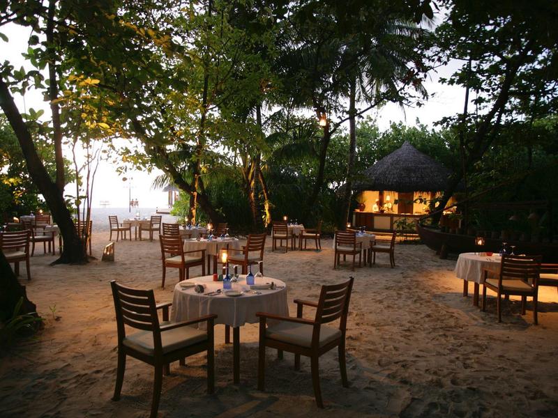Romantisches Strandrestaurant bei Sonnenuntergang mit gedeckten Tischen im Sand unter Bäumen.