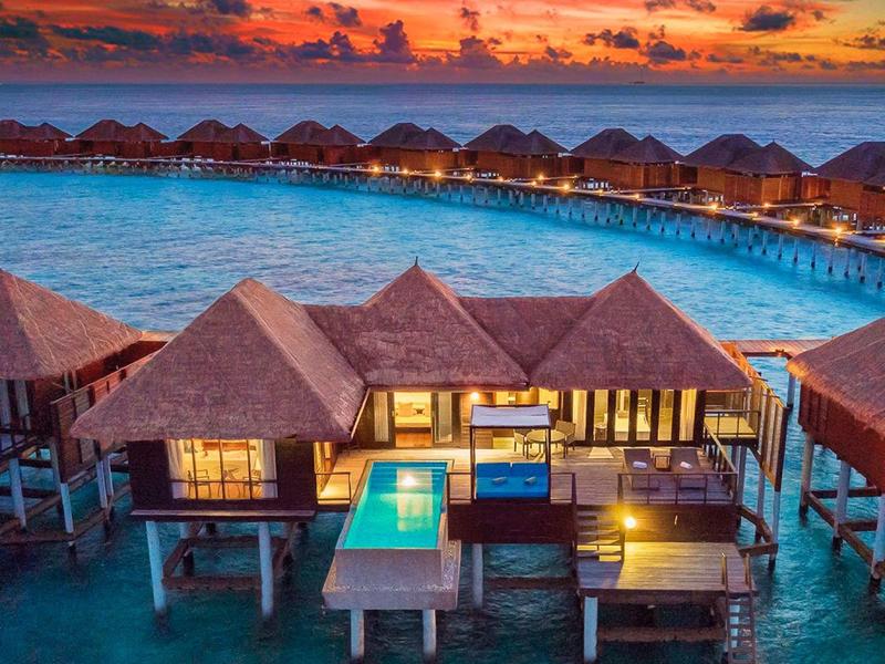 Bungalows auf Stelzen über klarem Wasser, darunter großer Pool, bunter Sonnenuntergang am Horizont.