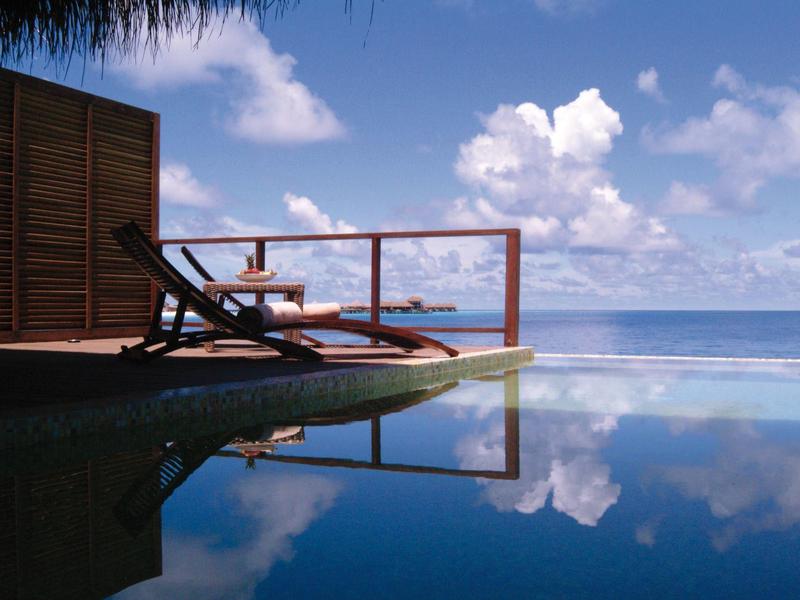 Pool mit Wasser, Liegestühle und blauer Himmel mit Wolken, Meer im Hintergrund, ruhige Atmosphäre.