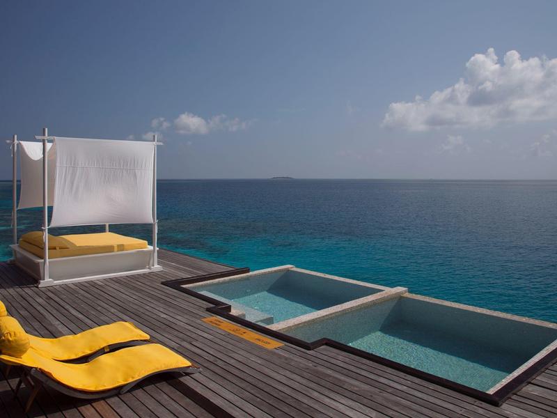 Terrasse mit Holzdeck, gelber Liege und weißem Baldachinbett neben zwei Pools am Meerufer.