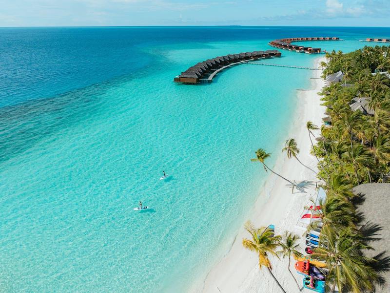 Türkisblaues Wasser mit Sandstrand und über Wasser gebauten Bungalows neben Palmen.
