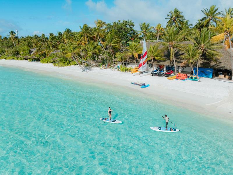 Strand mit weißem Sand, türkisfarbenem Wasser und zwei Menschen auf Paddleboards, umgeben von Palmen.
