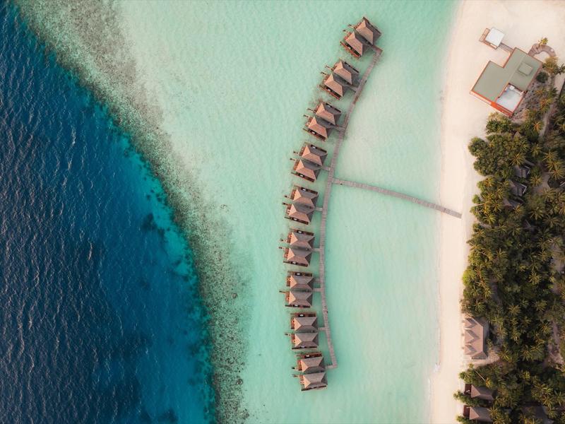 Luftaufnahme von Bungalows über klarem Wasser an einem weißen Sandstrand mit grüner Vegetation.