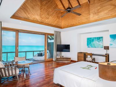 Helles Hotelzimmer mit Holzfußboden, Deckenventilator und Blick auf das blaue Meer durch große Fenster.