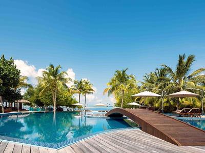 Rundlicher Hotelpool mit Holzbrücke, Sonnenschirmen und Palmen unter blauem Himmel.