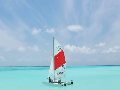 Kleines Segelboot mit rotem und weißem Segel auf klarem, türkisfarbenem Meer unter blauem Himmel.