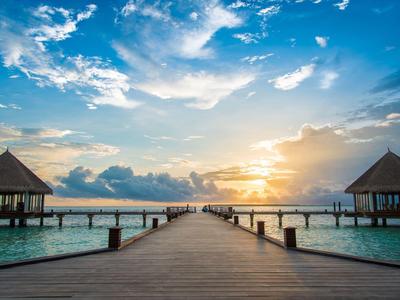 Langer Holzsteg führt zu zwei Hütten über türkisfarbenem Meer bei Sonnenuntergang.
