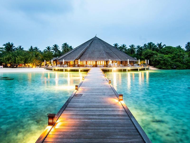 Ein hölzerner Steg führt zu einer beleuchteten Overwater-Villa bei Sonnenuntergang im türkisfarbenen Meer.