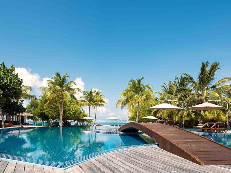 Rundlicher Hotelpool mit Holzbrücke, Sonnenschirmen und Palmen unter blauem Himmel.