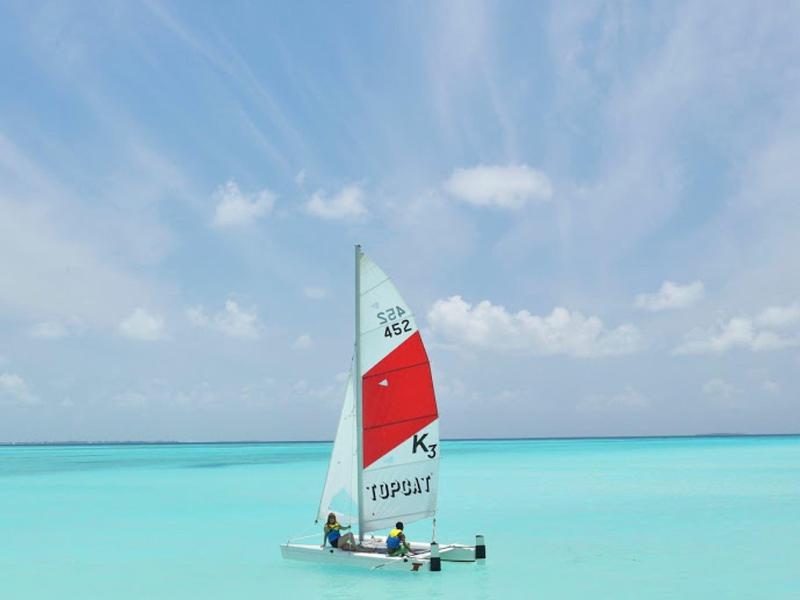 Ein kleines Segelboot mit weißem und rotem Segel auf türkisblauem, klarem Wasser unter blauem Himmel.