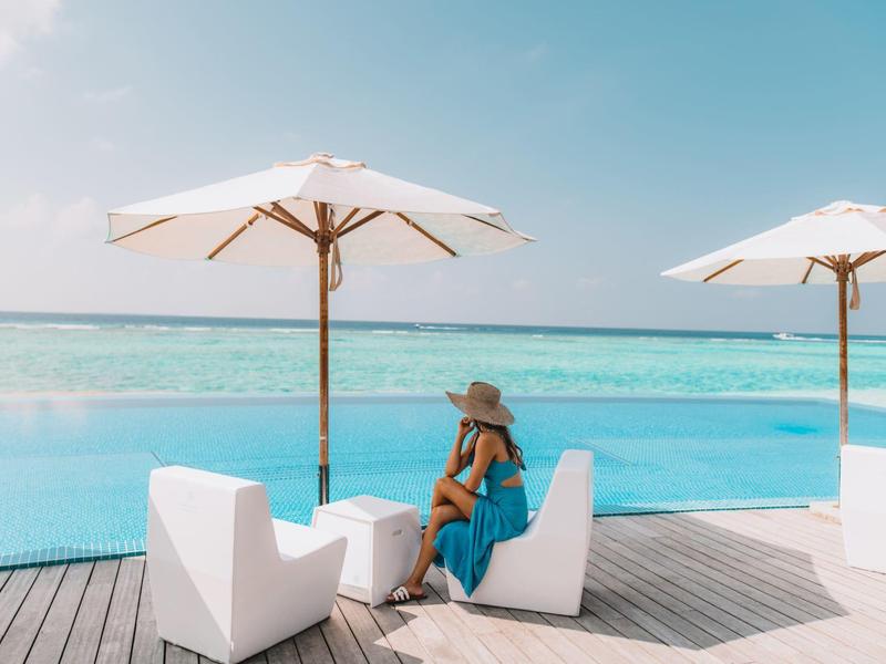 Frau in blauem Kleid sitzt unter Sonnenschirm am Pool mit Meerblick.