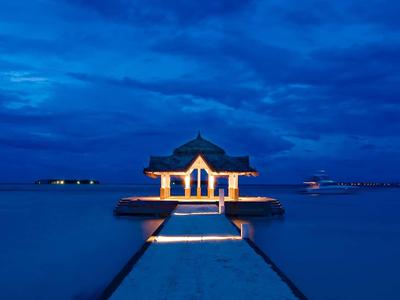 Ein beleuchteter Pavillon am Ende eines Stegs über ruhigem Wasser bei blauem Nachthimmel.